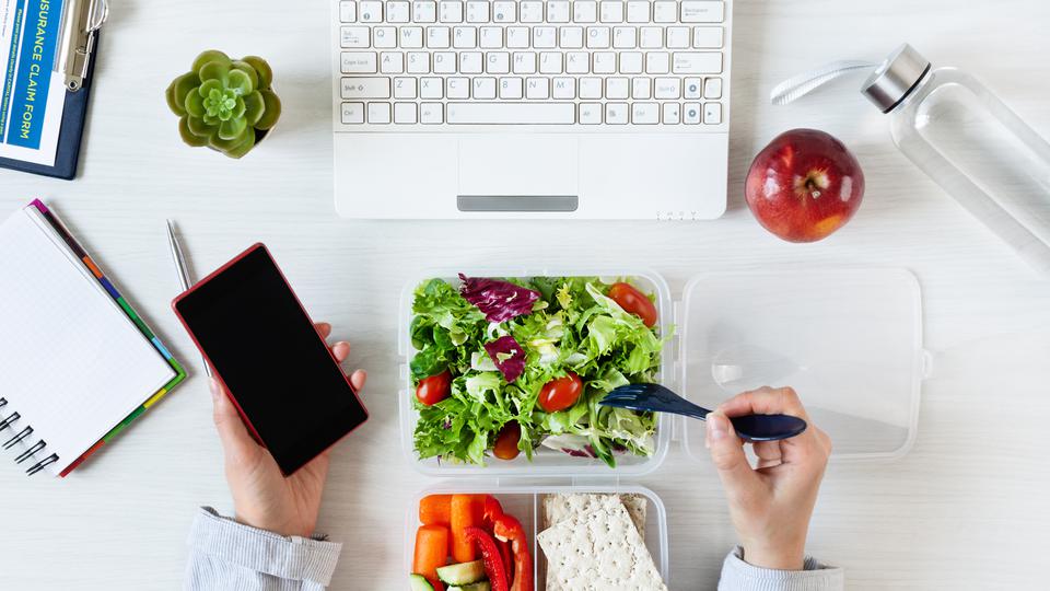 En person sitter vid en dator och äter mat från en matlåda