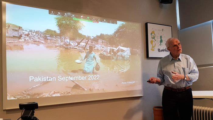 En äldre man talar framför en bildduk där man ser en man som vadar genom översvämmad mark. I bilden ligger texten Pakistan september 2022.  